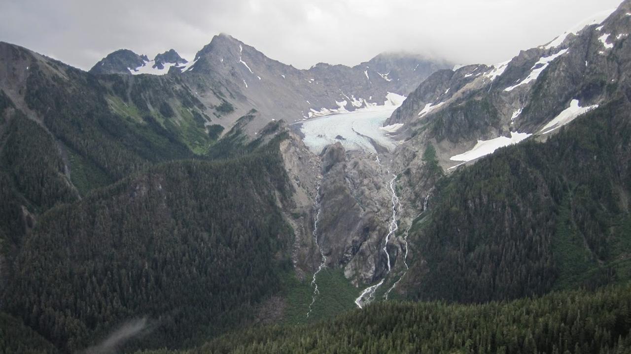 Der Schnee auf einem Gletscher zwischen zwei hohen Bergen franst nach unten ins Tal hinein aus und endet in einer Grünfläche.