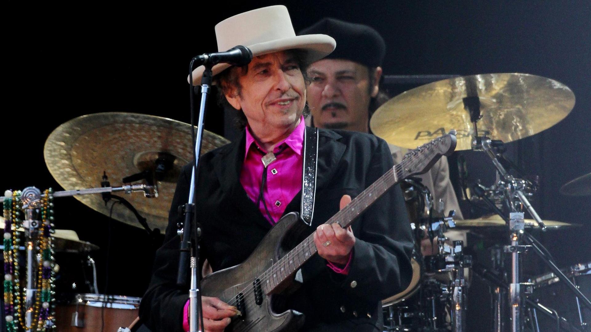 Der Sänger Bob Dylan bei einem Auftritt. Er trägt einen hellen Hut und ein pinkfarbenes Hemd. Der Sänger Bob Dylan bei einem Auftritt. Er trägt einen hellen Hut und ein pinkfarbenes Hemd.