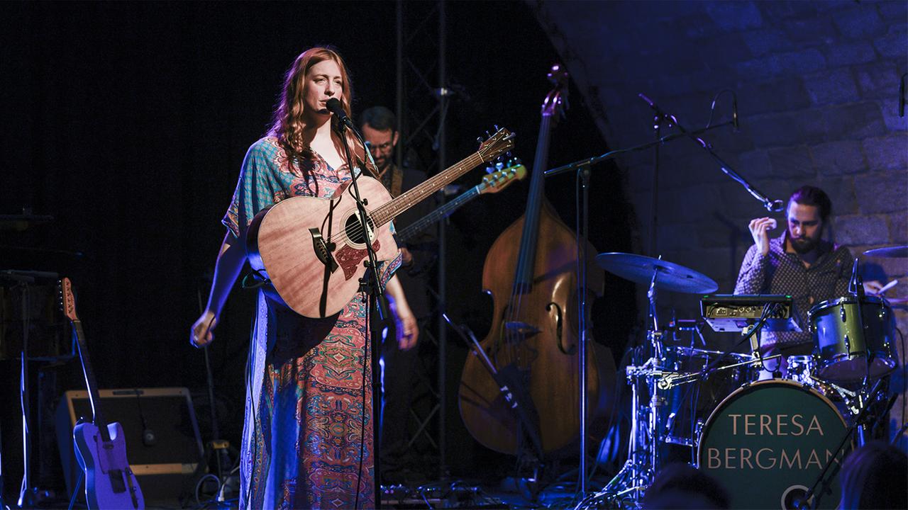 Eine Frau mit roten Locken steht in buntem Kleid auf der Bühne, eine Akustikgitarre vor sich. Hinter ihr Bassist und Schlagzeuger, sowie Kontrabass, E-Gitarre und Verstärker. Eine Frau mit roten Locken steht in buntem Kleid auf der Bühne, eine Akustikgitarre vor sich. Hinter ihr Bassist und Schlagzeuger, sowie Kontrabass, E-Gitarre und Verstärker.