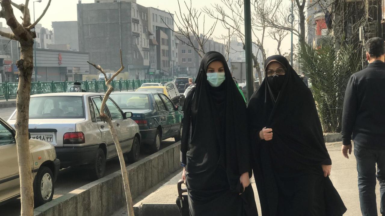 Zwei komplett in schwarzen langen Gewändern und mit Masken verhüllte Frauen gehen mit Einkaufswagen die Straße entlang. Zwei komplett in schwarzen langen Gewändern und mit Masken verhüllte Frauen gehen mit Einkaufswagen die Straße entlang.