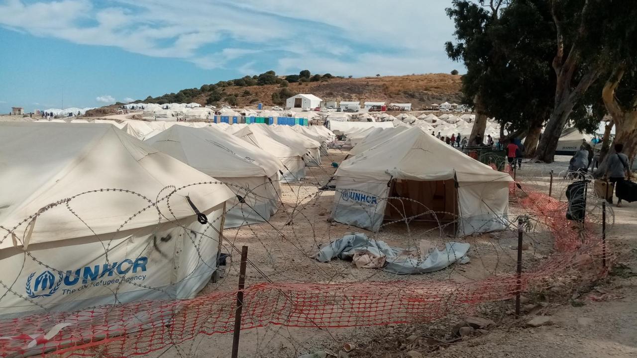 Mehrere weiße spitze Zelte des UNHCR stehen hinter einem Stacheldrahtzaun. Eines ist geöffnet, das Innere ist leer. Mehrere weiße spitze Zelte des UNHCR stehen hinter einem Stacheldrahtzaun. Eines ist geöffnet, das Innere ist leer.