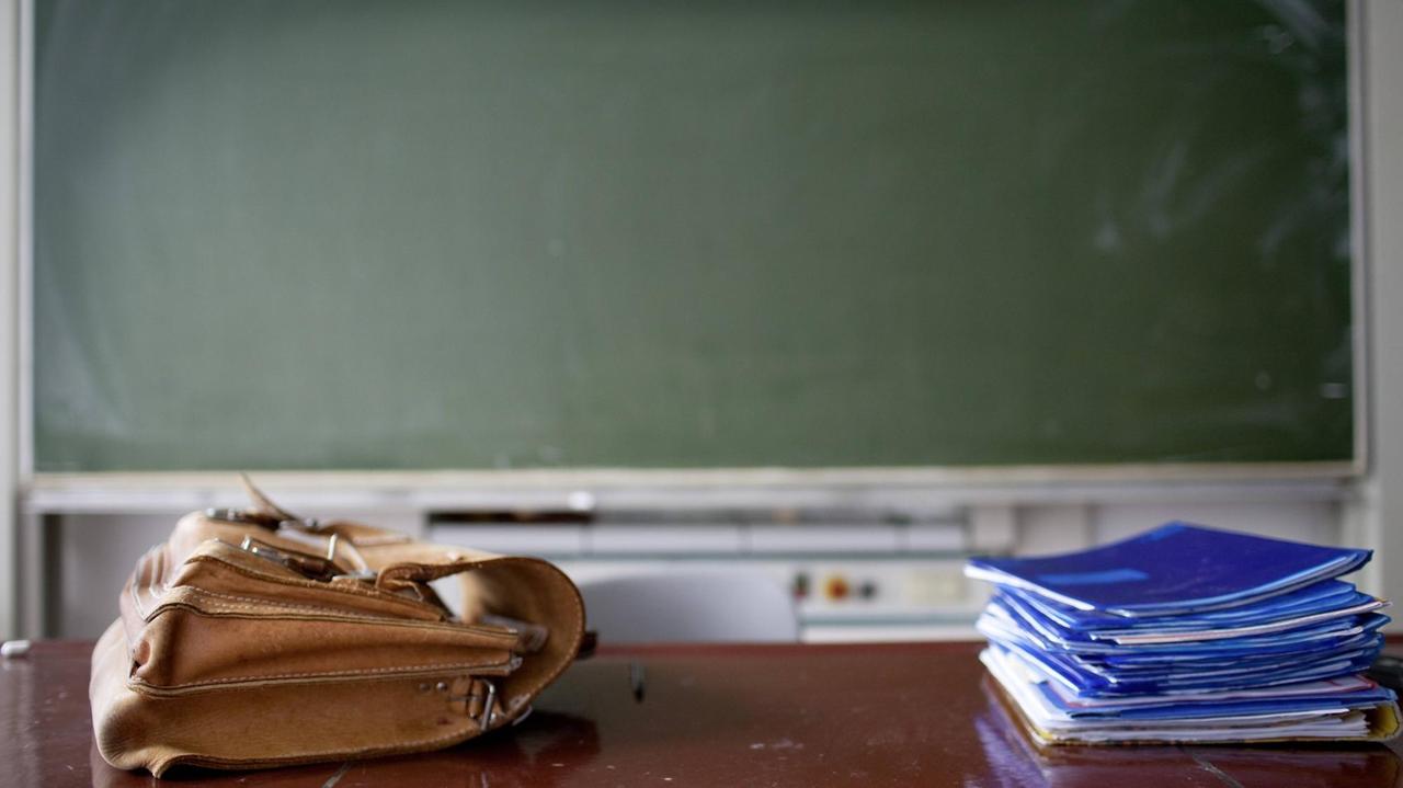 In einem Klassenzimmer liegen eine Aktentasche und ein Heftestapel auf dem Lehrerpult In einem Klassenzimmer liegen eine Aktentasche und ein Heftestapel auf dem Lehrerpult