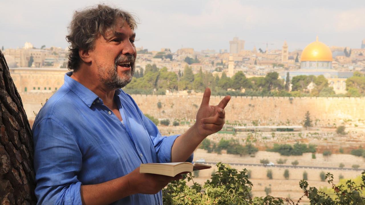 Der Passionsspiele-Regisseur Christian Stückl hält eine Bibel in der Hand. Im Hintergrund sieht man die goldene Kuppel des Felsendoms.