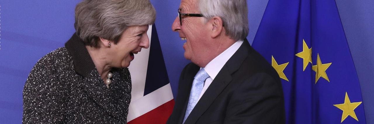 Theresa May und Jean-Claude Juncker halten ihre Hände gegenseitig. Sie lachen herzlich.
