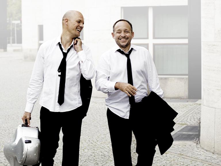 Zwei Männer in weißem Hemd und loser schwarzer Krawatte gehen lachend an einem Gebäude vorbei.