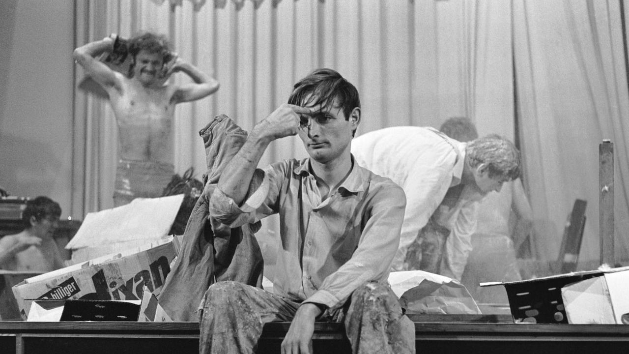 Ein junger Mann sitzt im Rahmen eines Happenings in einer Art Schlafanzug auf einem Tisch und hält die Hand an den Kopf.