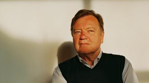 Joachim Lottmann sitzt auf einem Stuhl, er trägt Hemd und Pullunder. Im Hintergrund Schattenspiele.