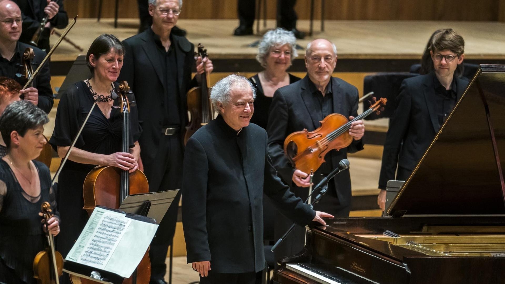 In einem Konzertsaal steht in der Bildmitte ein älterer Mann mit grauen Haaren, der sich mit der linken Hand an einem Flügel festhält. Um ihn stehen Orchestermusiker mit ihren Instrumenten. In einem Konzertsaal steht in der Bildmitte ein älterer Mann mit grauen Haaren, der sich mit der linken Hand an einem Flügel festhält. Um ihn stehen Orchestermusiker mit ihren Instrumenten.