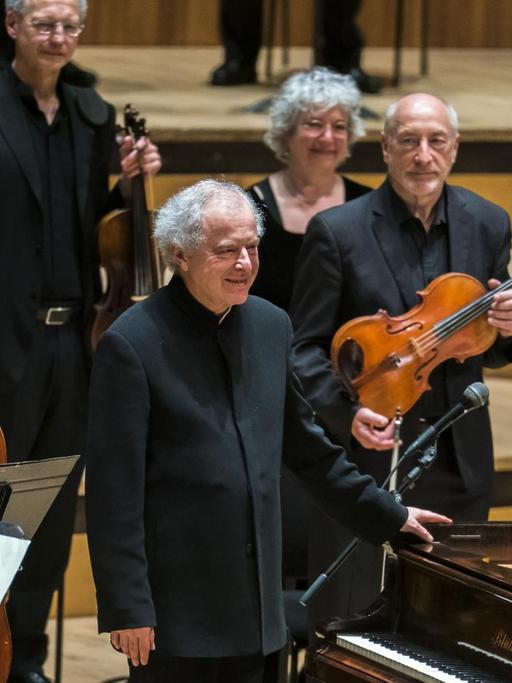 In einem Konzertsaal steht in der Bildmitte ein älterer Mann mit grauen Haaren, der sich mit der linken Hand an einem Flügel festhält. Um ihn stehen Orchestermusiker mit ihren Instrumenten.