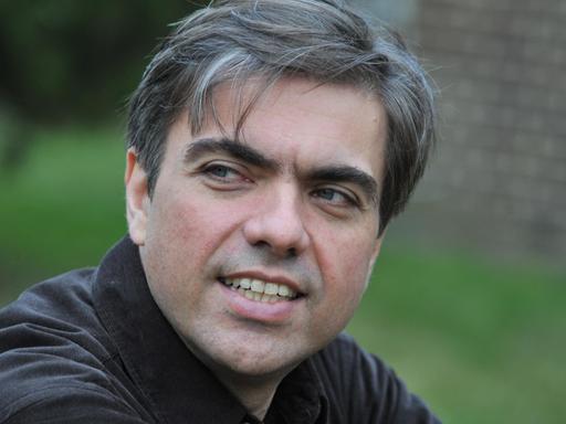 Ein Mann mit dunklen und grauen Haaren blickt nach links aus dem Bild, im Hintergrund ist verschwommen Grün zu erkennen.