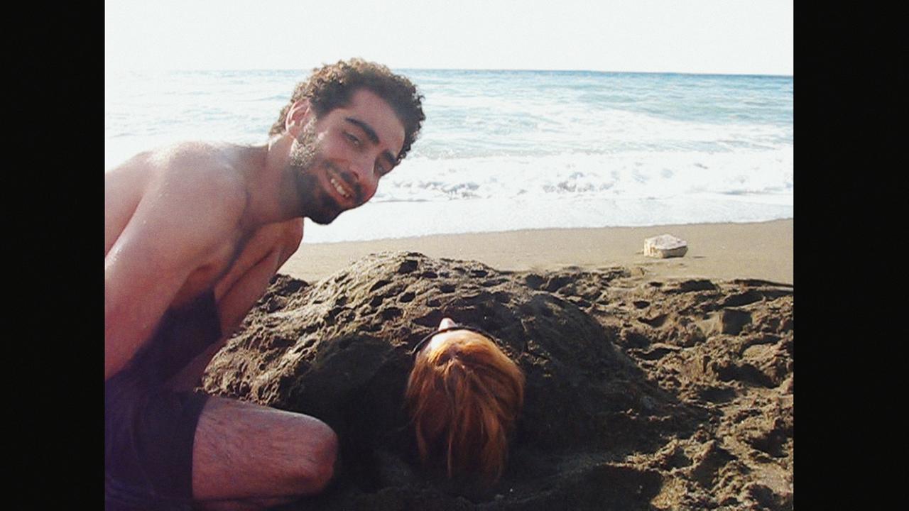 Ausschnitt aus dem Dokumentatfilm "The War Show": Ein junger Syrer am Strand beugt sich lachend über eine im Sand bis auf den Kopf eingebuddelte Frau Ausschnitt aus dem Dokumentatfilm "The War Show": Ein junger Syrer am Strand beugt sich lachend über eine im Sand bis auf den Kopf eingebuddelte Frau