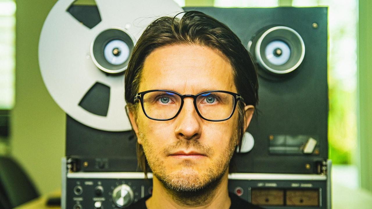 Ein Mann mit schwarzer Brille, T-Shirt und dunklen Haaren steht vor einem Tonbandgerät und blickt in die Kamera. Ein Mann mit schwarzer Brille, T-Shirt und dunklen Haaren steht vor einem Tonbandgerät und blickt in die Kamera.