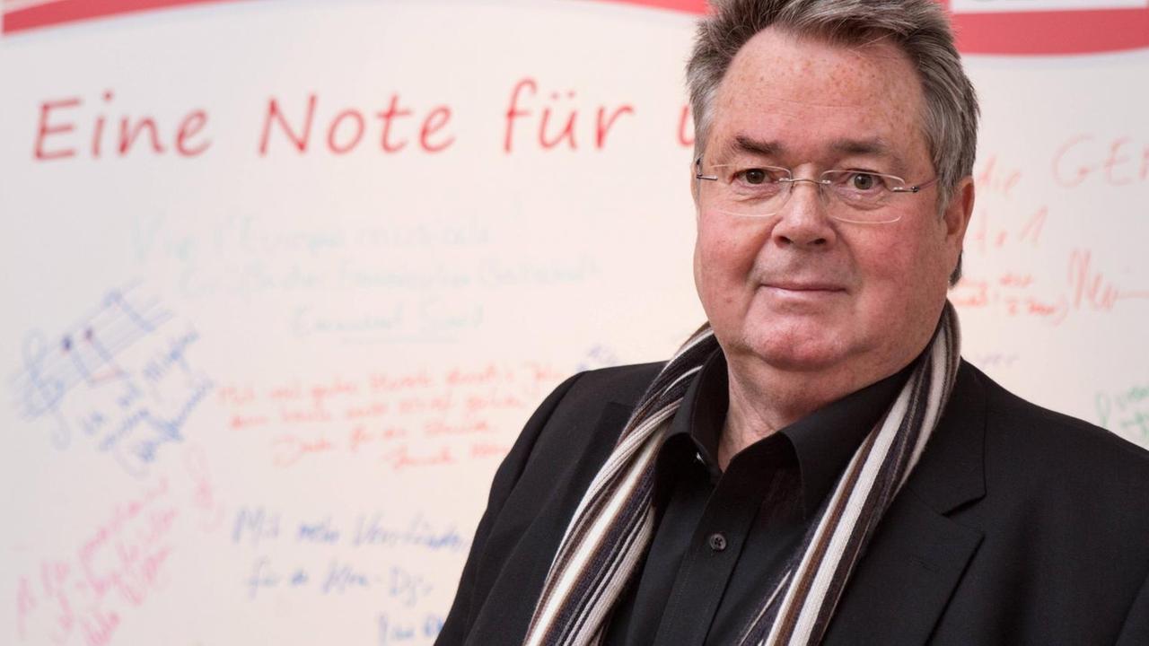 Ein grauhaariger Mann trägt schwarzes Jacket, um den Hals einen gestreiften Schal und blickt durch seine randlose Brille in die Kamera. Ein grauhaariger Mann trägt schwarzes Jacket, um den Hals einen gestreiften Schal und blickt durch seine randlose Brille in die Kamera.