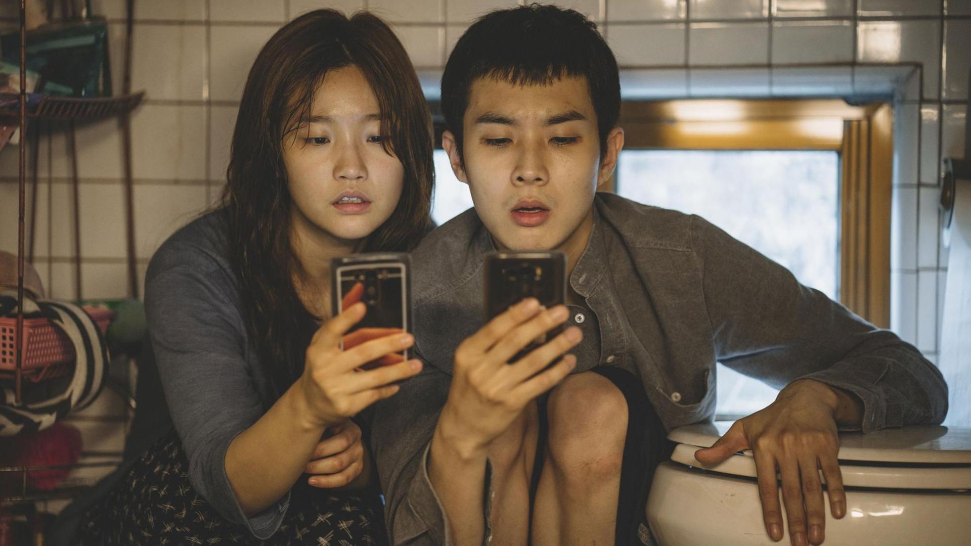 Szene aus dem Film "Parasite" von Regisseur Joon-ho Bong. Eine junge Frau und ein junger Mann sitzen in einem Badezimmer und starren auf ihr Smartphone. Szene aus dem Film "Parasite" von Regisseur Joon-ho Bong. Eine junge Frau und ein junger Mann sitzen in einem Badezimmer und starren auf ihr Smartphone.