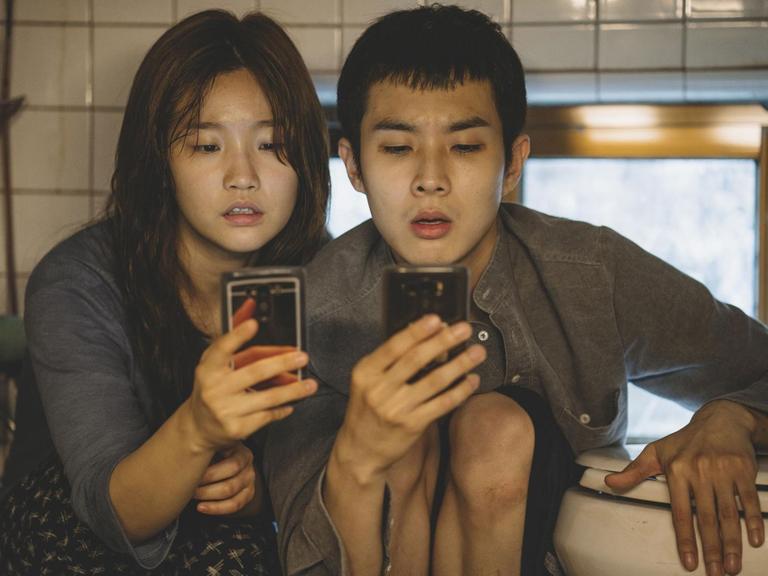 Szene aus dem Film "Parasite" von Regisseur Joon-ho Bong. Eine junge Frau und ein junger Mann sitzen in einem Badezimmer und starren auf ihr Smartphone.