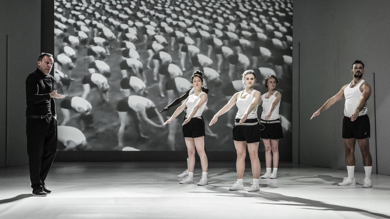 Auf einer Theaterbühne gibt ein schwarz gekleideter Mann anderen Schauspielern in Turnanzügen Anweisungen, dahinter eine Video-Leinwand mit Turnern in ebenfalls schwarz-weißen Turnanzügen. Auf einer Theaterbühne gibt ein schwarz gekleideter Mann anderen Schauspielern in Turnanzügen Anweisungen, dahinter eine Video-Leinwand mit Turnern in ebenfalls schwarz-weißen Turnanzügen.