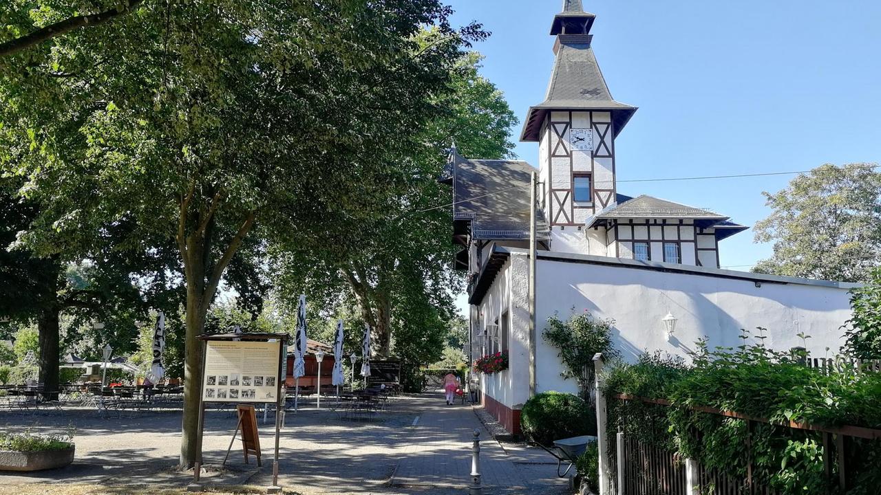 An einem Weg mit Bäumen liegt ein verspieltes weißes Gebäude aus dem 19. Jahrhundert mit einem Turm. An einem Weg mit Bäumen liegt ein verspieltes weißes Gebäude aus dem 19. Jahrhundert mit einem Turm.