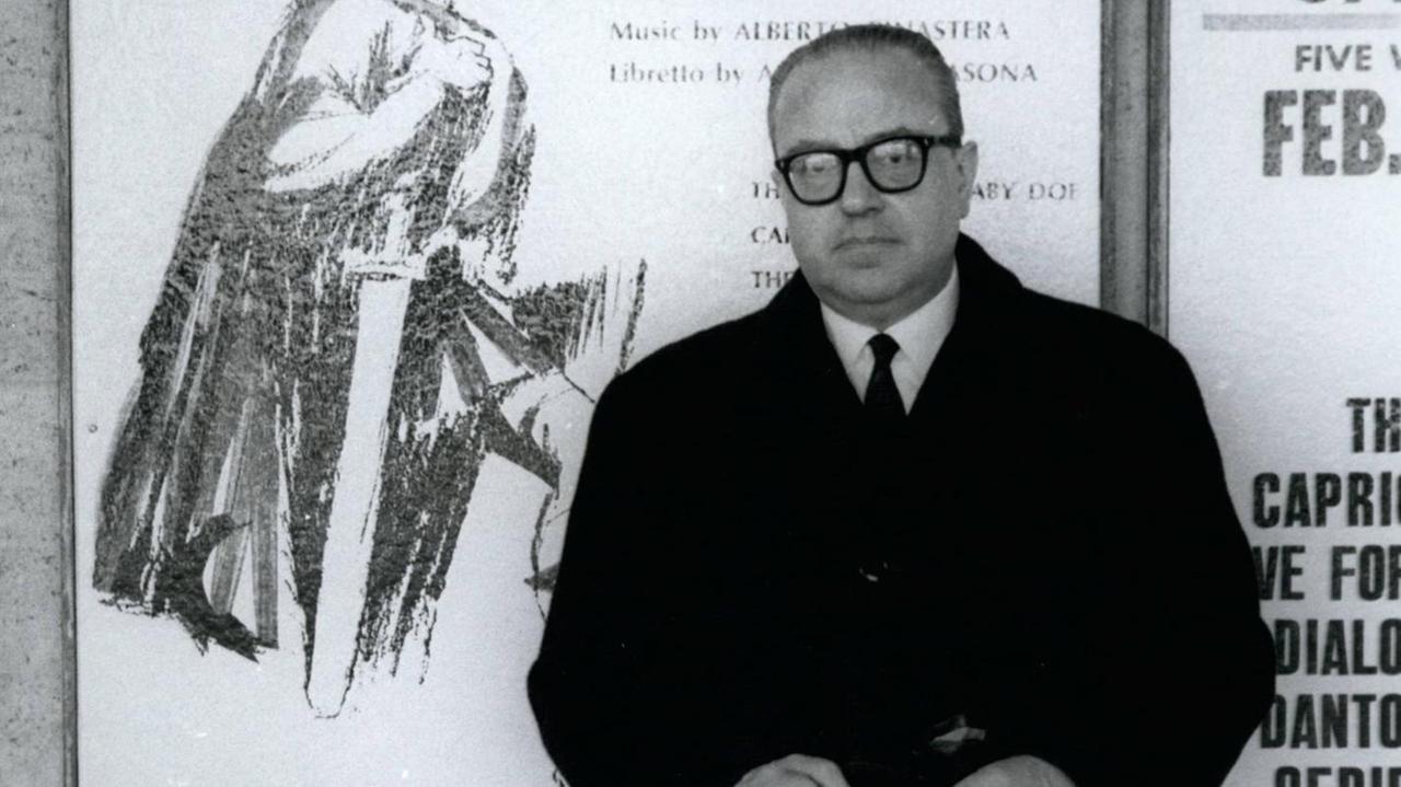 Der argentinische Komponist Alberto Ginastera steht vor einem Plakat mit einer Opern-Ankündigung. Schwarz-weiß-Bild. Der argentinische Komponist Alberto Ginastera steht vor einem Plakat mit einer Opern-Ankündigung. Schwarz-weiß-Bild.