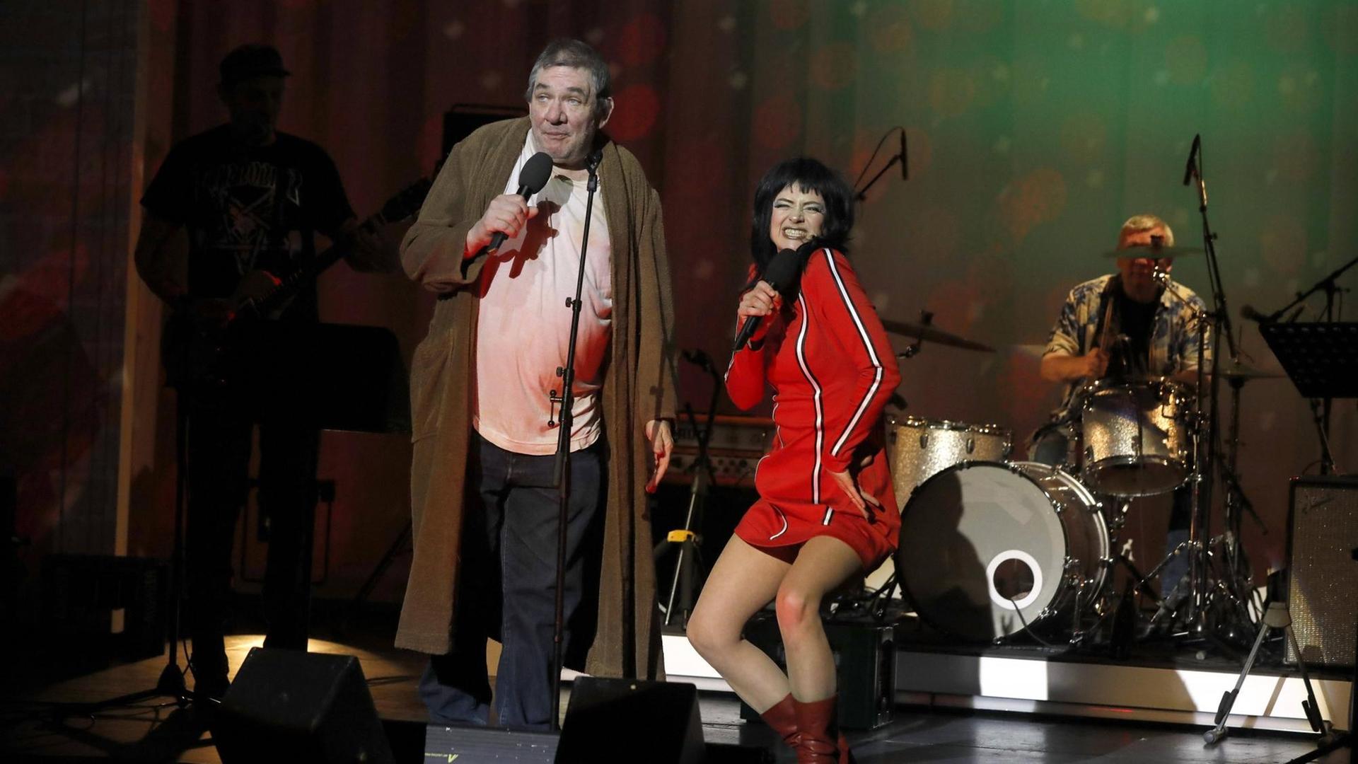 Ein Mann steht auf einer Bühne, er trägt einen Bademantel und hält ein Mikrofon in der Hand. Neben ihm steht eine Frau in einem roten Kleid.