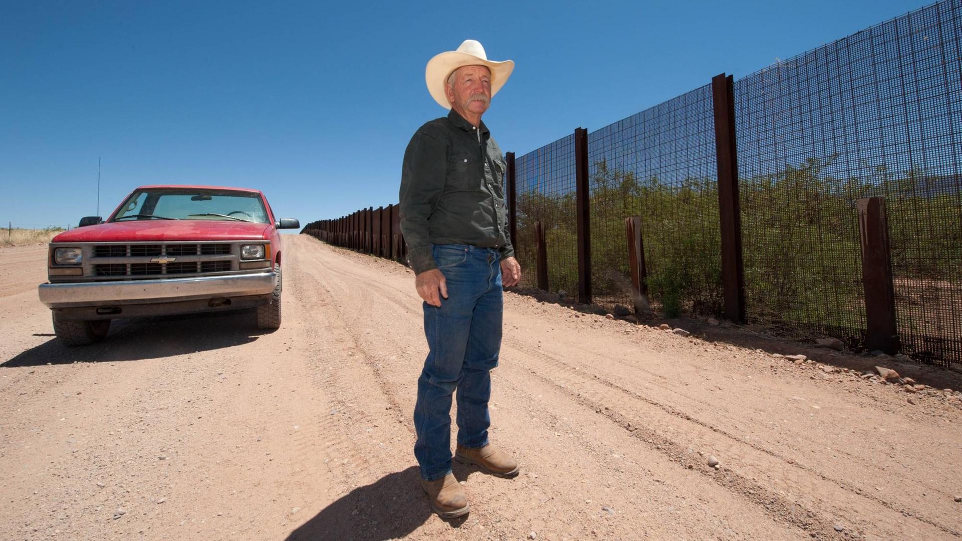 John Ladd steht auf einer Schotterstraße, rechts von ihm ein Grenzzaun, links hinter ihm sein roter verbeulter Pickup