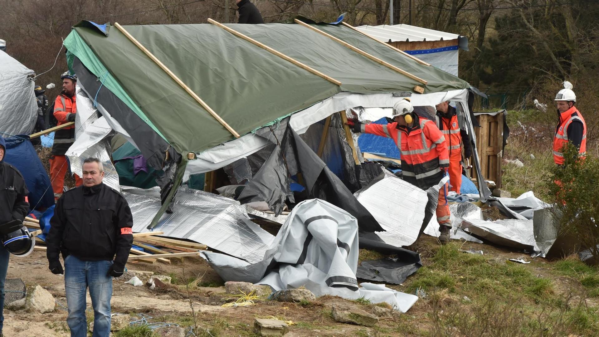 Arbeiter mit orangefarbenen Schutzwesten und Helmen machen sich einer selbsterrichteten Baracke aus Holz und Plastikplanenzu schaffen. Arbeiter mit orangefarbenen Schutzwesten und Helmen machen sich einer selbsterrichteten Baracke aus Holz und Plastikplanenzu schaffen.