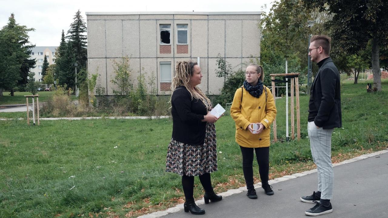 Eine junge Frau mit blonden Rastalocken im Kleid und ein junger Mann in Stoffhose und Jackett reden auf der Straße mit einer jungen Frau im gelben Regenmantel. Eine junge Frau mit blonden Rastalocken im Kleid und ein junger Mann in Stoffhose und Jackett reden auf der Straße mit einer jungen Frau im gelben Regenmantel.