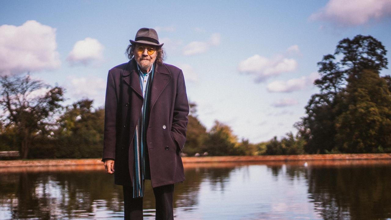 Bill Fay steht mit gelb getönter Sonnenbrille, Hut und Mantel vor einem Teich. Bill Fay steht mit gelb getönter Sonnenbrille, Hut und Mantel vor einem Teich.