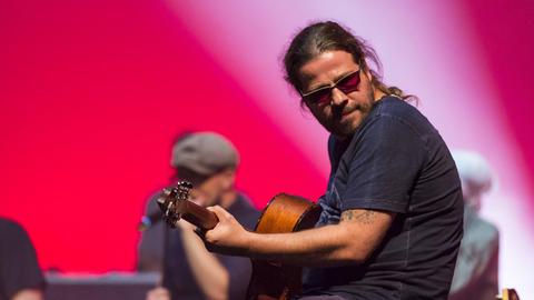 Gitarrist Andreas Bayless von der deutschen Band Söhne Mannheims live beim Blue Balls Festival im Konzertsaal des KKL Luzern, Schweiz. Der Musiker trägt eine Sonnenbrille und hat die Haare zum Zopf zusammengebunden.