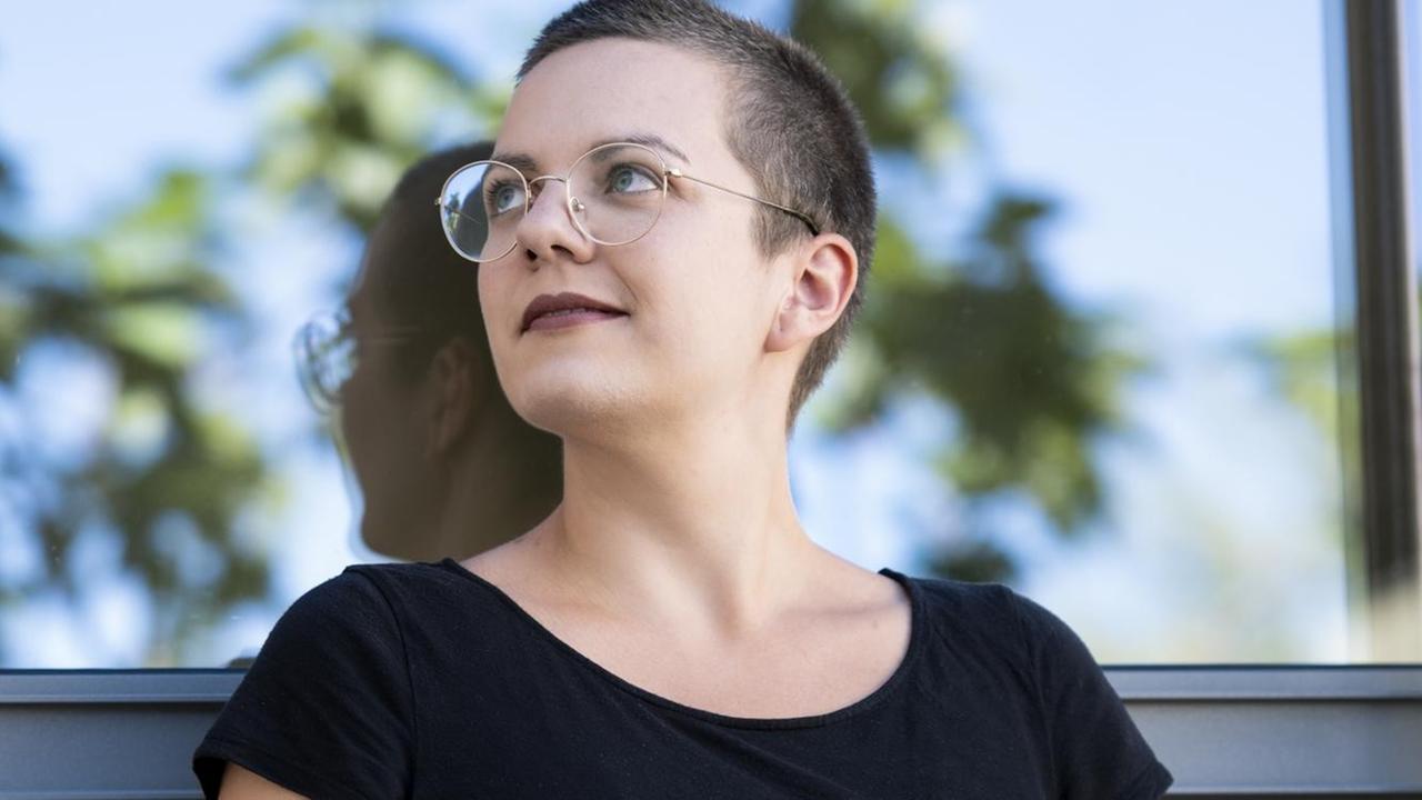 Eine junge Frau mit kurzen Haare und runder Brille lehnt an einer Fensterwand und schaut schräg nach oben. Eine junge Frau mit kurzen Haare und runder Brille lehnt an einer Fensterwand und schaut schräg nach oben.