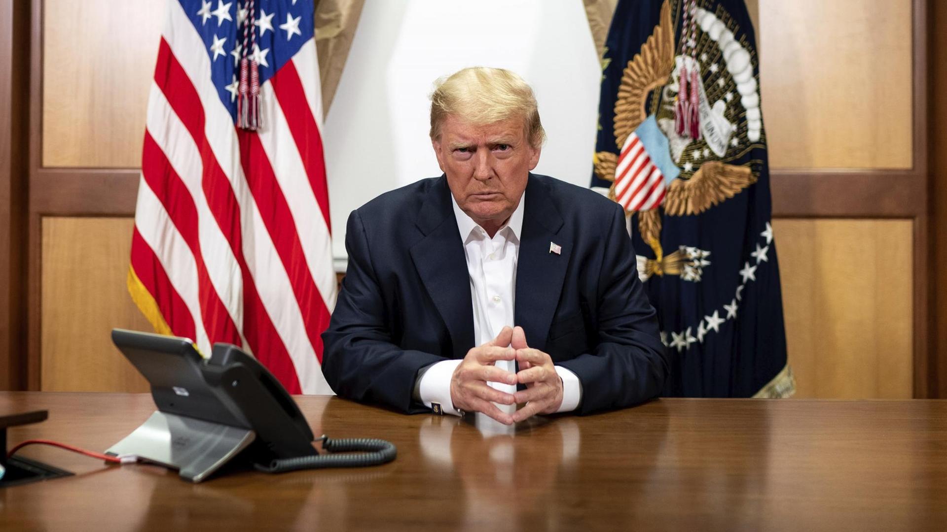 Trump sitzt vor der US-Fahne und der Präsidentenfahne mit konzentriertem Blick in die Kamera an einem Tisch. Trump sitzt vor der US-Fahne und der Präsidentenfahne mit konzentriertem Blick in die Kamera an einem Tisch.