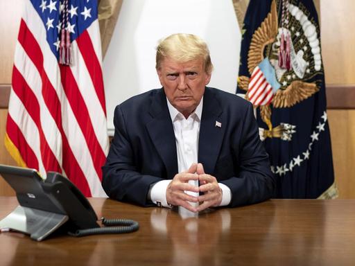 Trump sitzt vor der US-Fahne und der Präsidentenfahne mit konzentriertem Blick in die Kamera an einem Tisch.