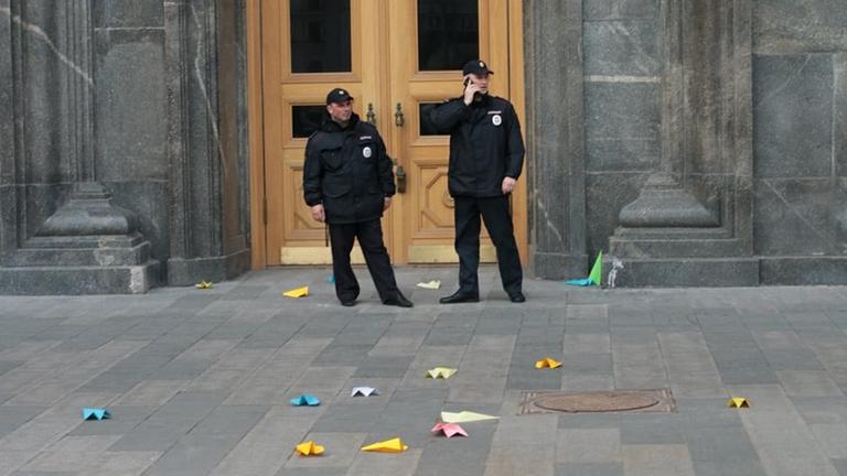 Bunte Papeirflieger liegen am Boden vor einem Gebäude, das von zwei Wachleuten bewacht wird. Bunte Papeirflieger liegen am Boden vor einem Gebäude, das von zwei Wachleuten bewacht wird.