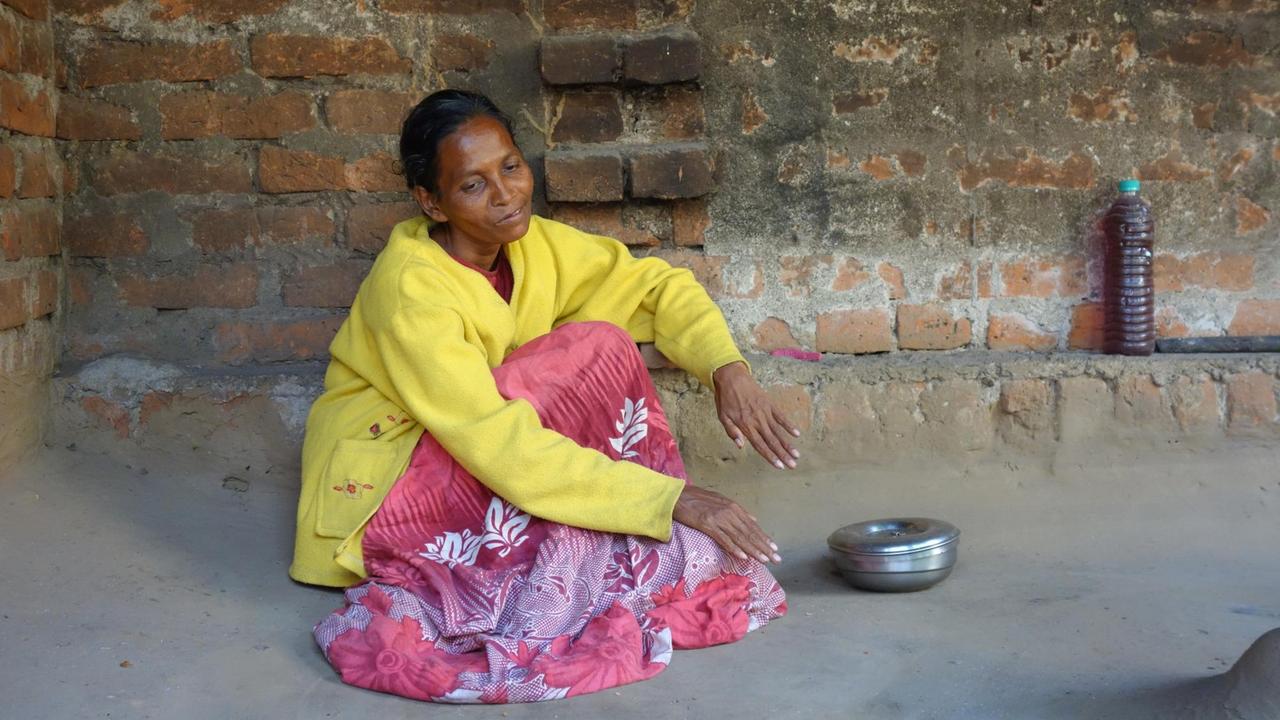 Eine magere Frau um die 40 in lachsrot-lila geblümtem Kleid und gelber Jacke sitzt auf dem Betonboden einer Terrasse.