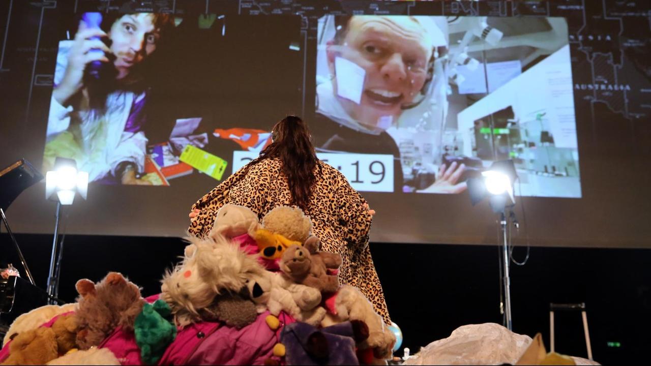Eîne Frau mit Leopardenmantel steht mit dem Rücken zur Kamera, hinter ihr ein Kuscheltierberg, vor ihr eine Leinwand mit Videokonferenz. Eîne Frau mit Leopardenmantel steht mit dem Rücken zur Kamera, hinter ihr ein Kuscheltierberg, vor ihr eine Leinwand mit Videokonferenz.