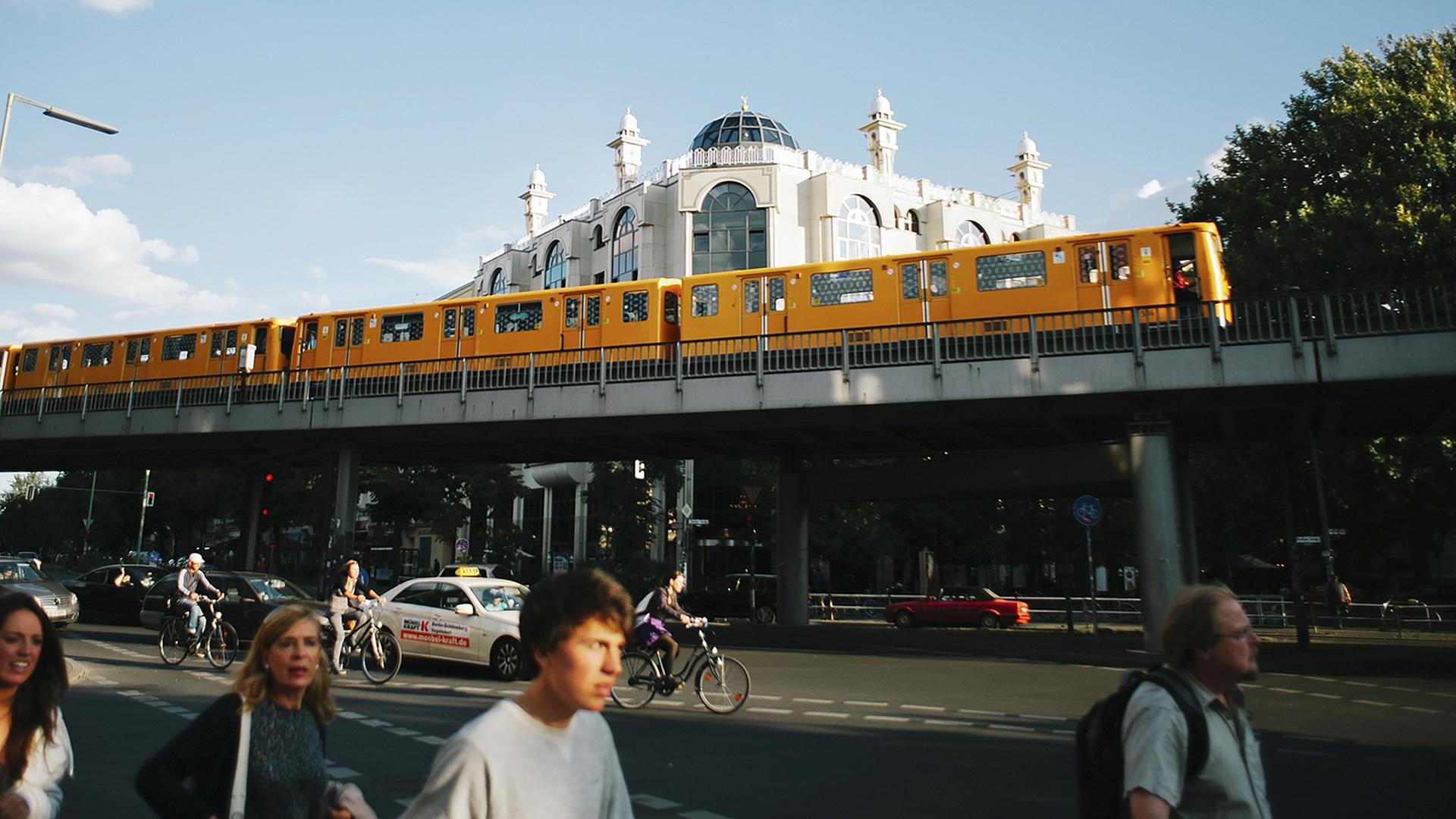 Im Vordergrund fährt die typisch gelbe Berliner U-Bahn, dahinter ist die Omar-ibn-Al-Khattab Moschee, ein weißes Eckgebäude, zu sehen.