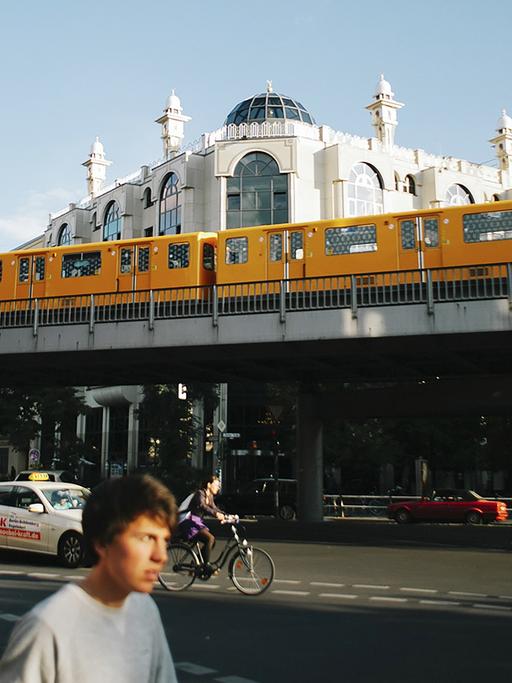 Im Vordergrund fährt die typisch gelbe Berliner U-Bahn, dahinter ist die Omar-ibn-Al-Khattab Moschee, ein weißes Eckgebäude, zu sehen.