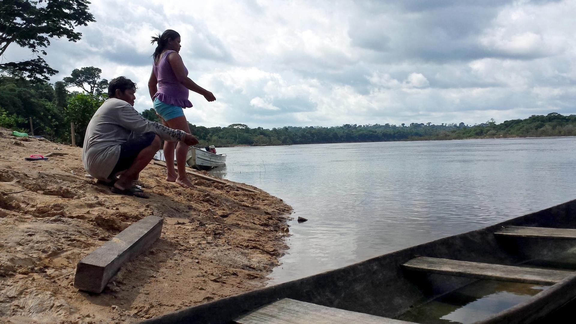 Ein Mann und eine Frau indianischen Ursprungs am Flussufer eines weiten Stroms, im Vordergrund ein Boot