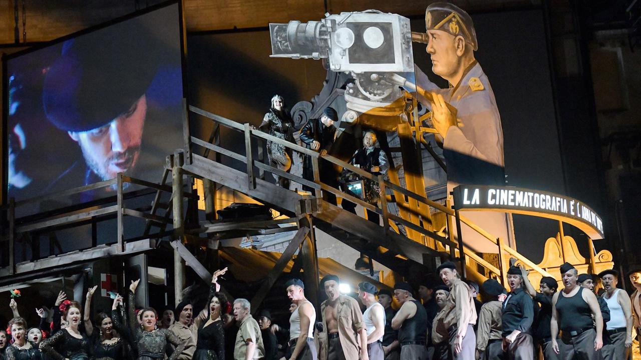 Zu sehen ist eine große Leinwand, auf die ein Mann projiziert wird, ein Chor, eine Treppe und eine überdimensionierte Nachbildung eines Offiziers, der durch eine Kamera blickt. Im Hintergrund befindet sich ein Gebäude mit der Aufschrift "La Cinematografie".