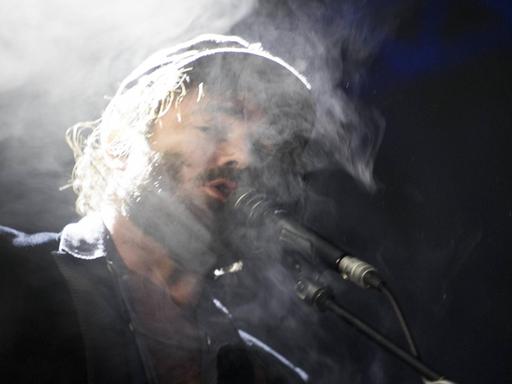 Der australische Sänger Angus Stone steht auf einer Bühne und singt in ein Mikrofon, um seinen Kopf ist Dampf.