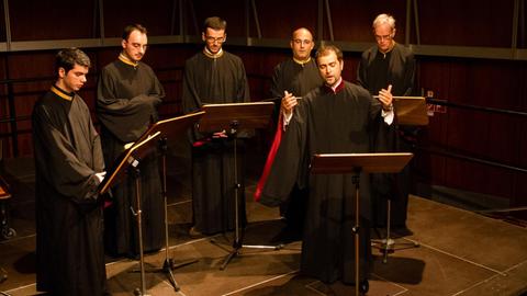 Sechs Männer in Roben stehen vor Notenständern auf einer Bühne.