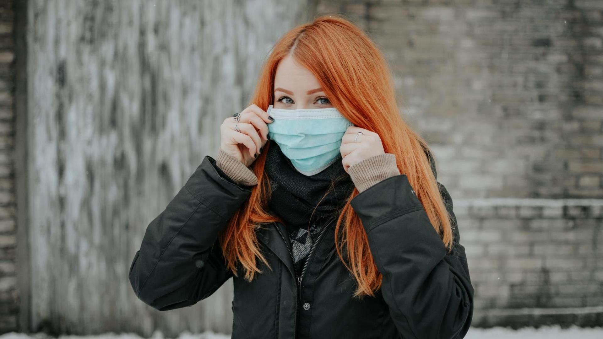 Eine rothaarige Frau in Winterjacke trägt eine Schutzmaske und schaut skeptisch. Eine rothaarige Frau in Winterjacke trägt eine Schutzmaske und schaut skeptisch.
