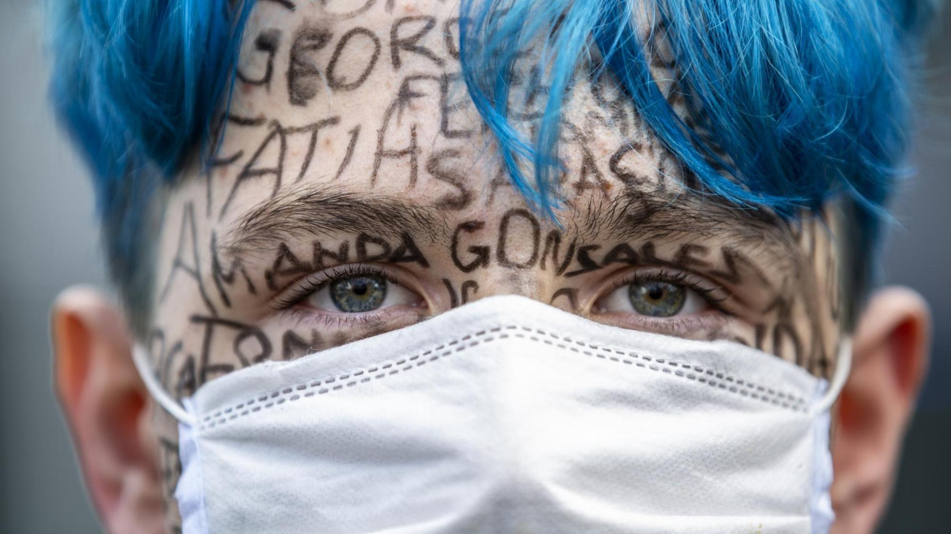 Ein Mann protestiert gegen Rassismus und Polizeibrutalität am 06. Juni 2020 auf dem Alexanderplatz in Berlin, Deutschland. Er trägt einen weißen Mundschutz und hat blau gefärbte Haare. Auf sein Gesicht sind verschiedene Namen mit Filzstift geschrieben.