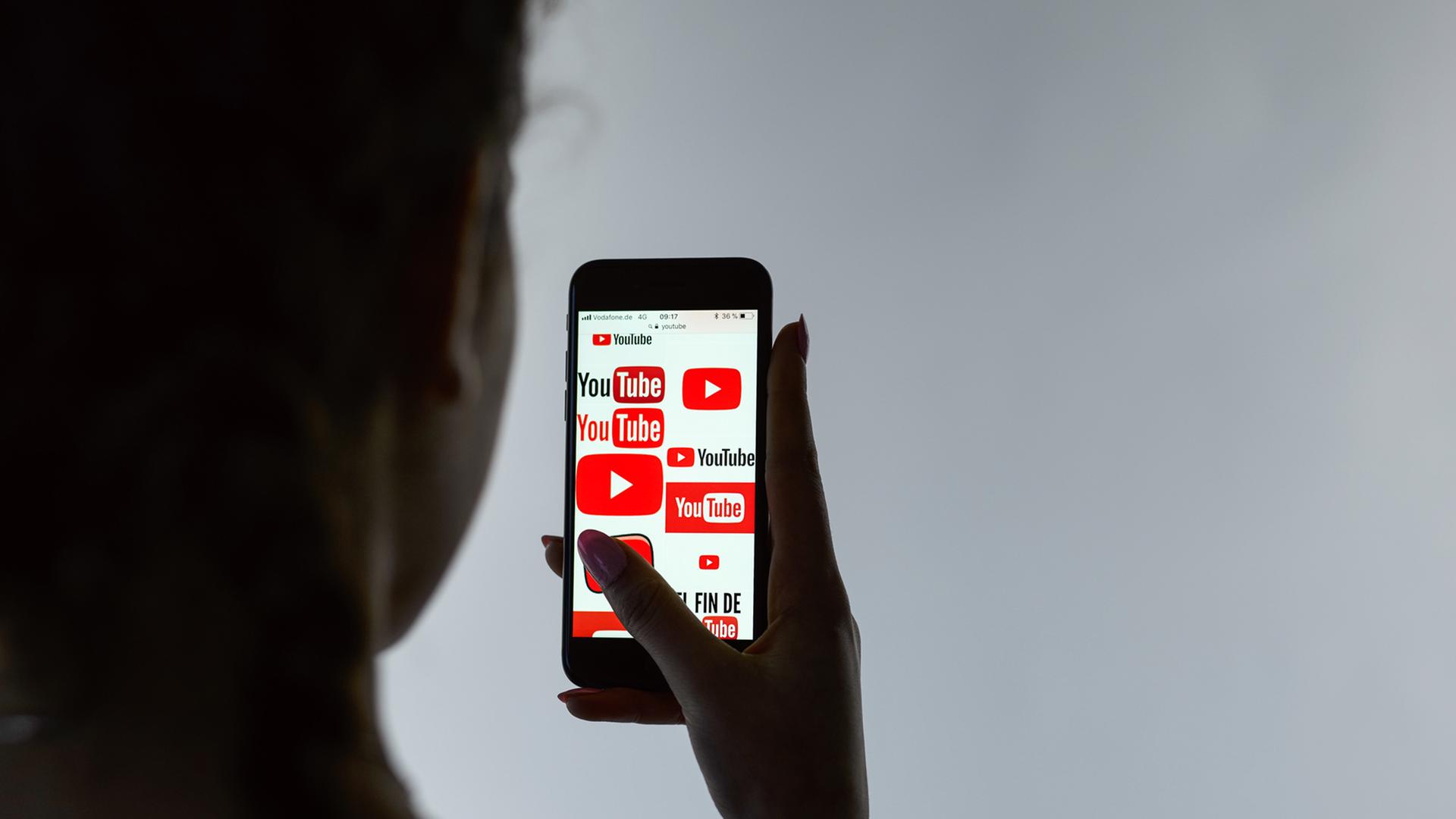 Wir schauen einer Frau über die Schulter, die ein Mobiltelefon hält, auf dem Logos von YouTube zu sehen sind. Wir schauen einer Frau über die Schulter, die ein Mobiltelefon hält, auf dem Logos von YouTube zu sehen sind.