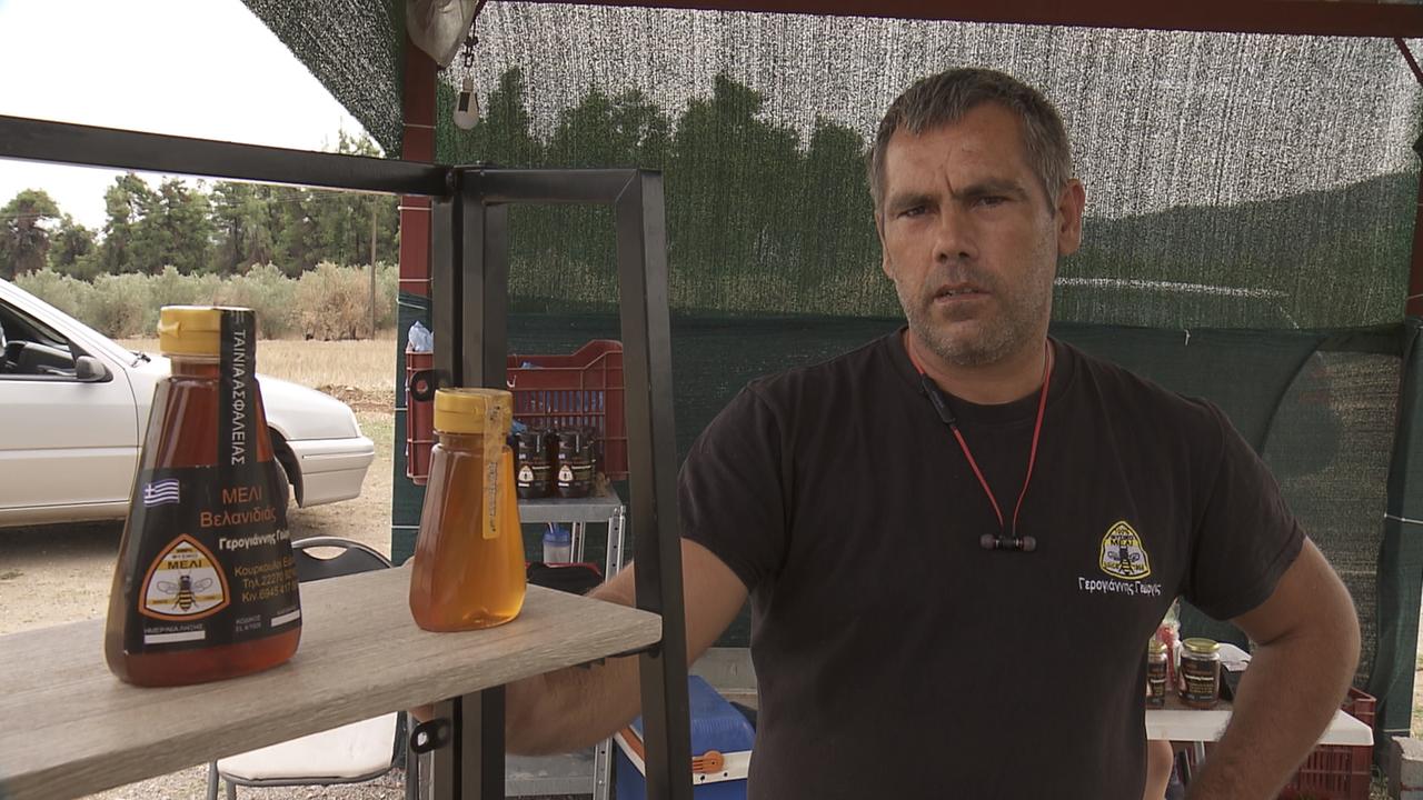 Ein Mann mit kritischem Blick im braunen T-Shirt und Head-Set um den Hals steht an einem Stand auf dem Honiglaschen mit Etiketten stehen. Ein Mann mit kritischem Blick im braunen T-Shirt und Head-Set um den Hals steht an einem Stand auf dem Honiglaschen mit Etiketten stehen.