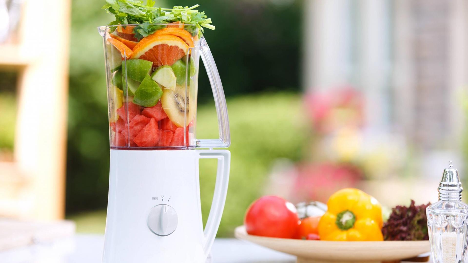 Ein Standmixer steht gefüllt mit verschiedenem Obst und Gemüse neben einem Teller mit Tomaten, Paprika und Salat. Ein Standmixer steht gefüllt mit verschiedenem Obst und Gemüse neben einem Teller mit Tomaten, Paprika und Salat.