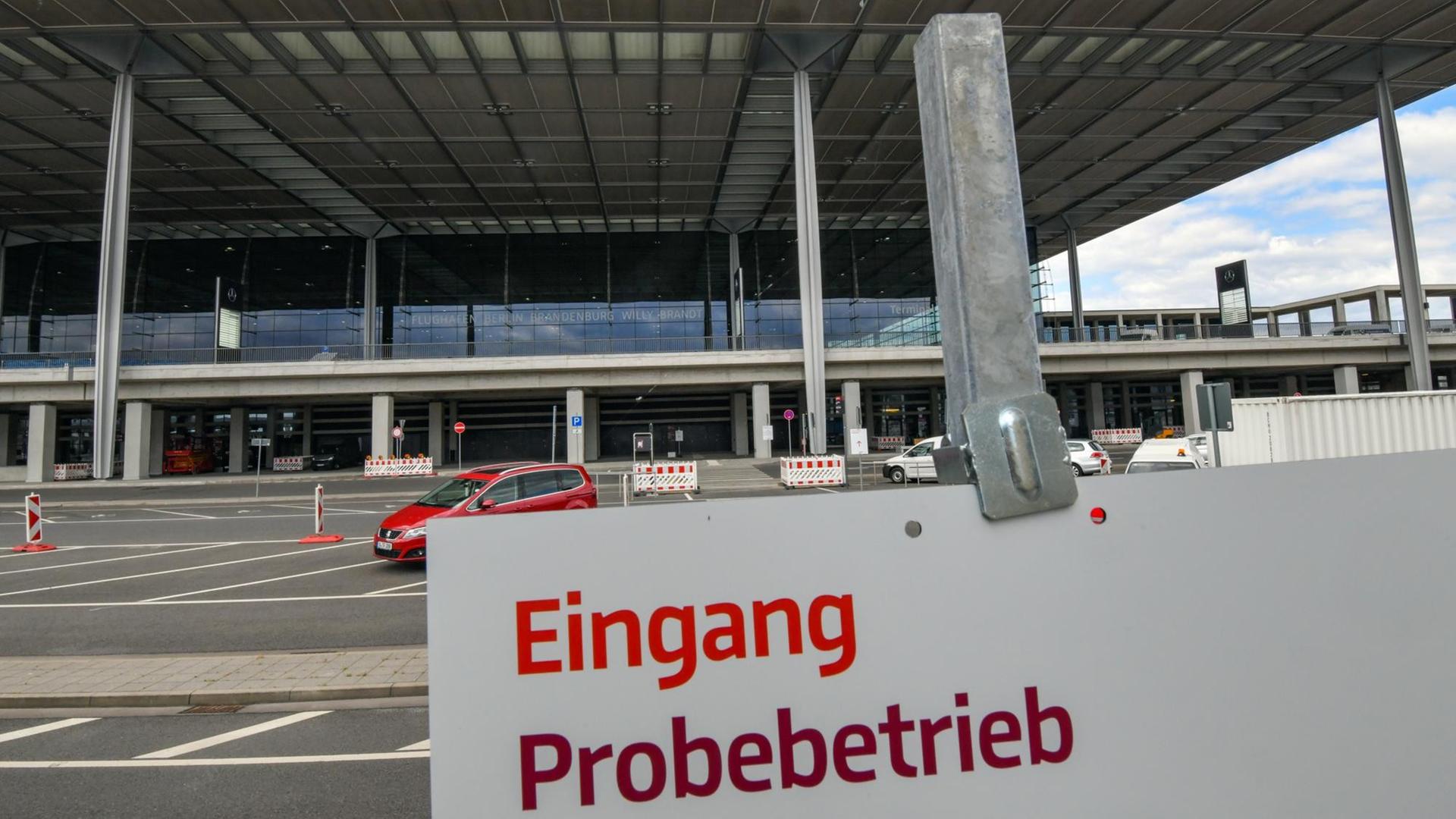 Das weiße Schild mit roter Schrift ist im Anschnitt im Vordergrund zu sehen. Dahinter ein Terminal von außen, vor dem wenige Autos stehen. Das weiße Schild mit roter Schrift ist im Anschnitt im Vordergrund zu sehen. Dahinter ein Terminal von außen, vor dem wenige Autos stehen.