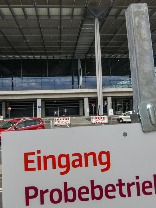 Das weiße Schild mit roter Schrift ist im Anschnitt im Vordergrund zu sehen. Dahinter ein Terminal von außen, vor dem wenige Autos stehen.