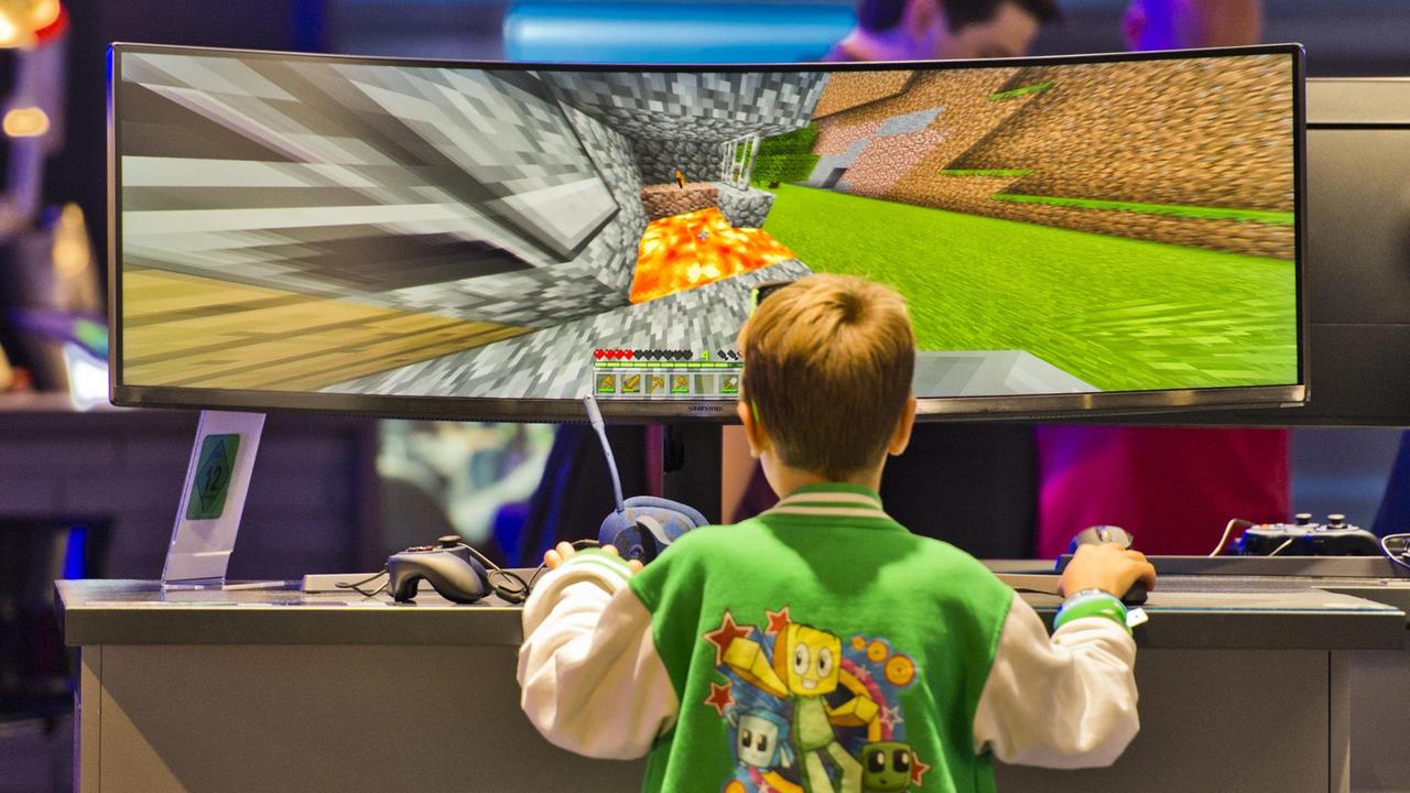 
Ein Junge steht vor einem Widescreen Monitor. Man sieht ihn von hinten wie er ein Spiel ausprobiert. Der Monitor ist im Verhältnis zu seinem Kopf riesig.