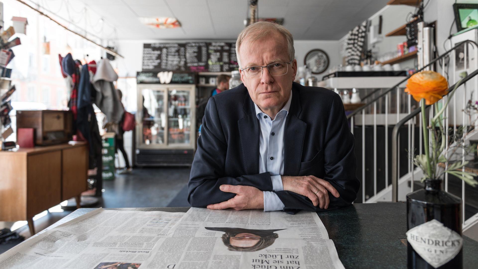Der Lyriker Henning Ziebritzki sitzt in einem Café, vor sich auf dem Tisch eine aufgeschlagene Zeitung.