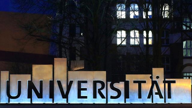 Außenansicht eines Universitätsgebäudes bei Nacht mit hellerleuchteten Fenstern, im Vordergrund der Schriftzug "Universität".